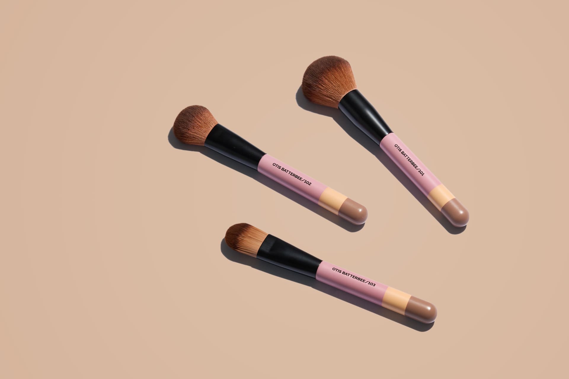 Three Otis Batterbee makeup brushes with pink handles on a beige back background. Foundation, Powder and Blusher Makeup Brushes