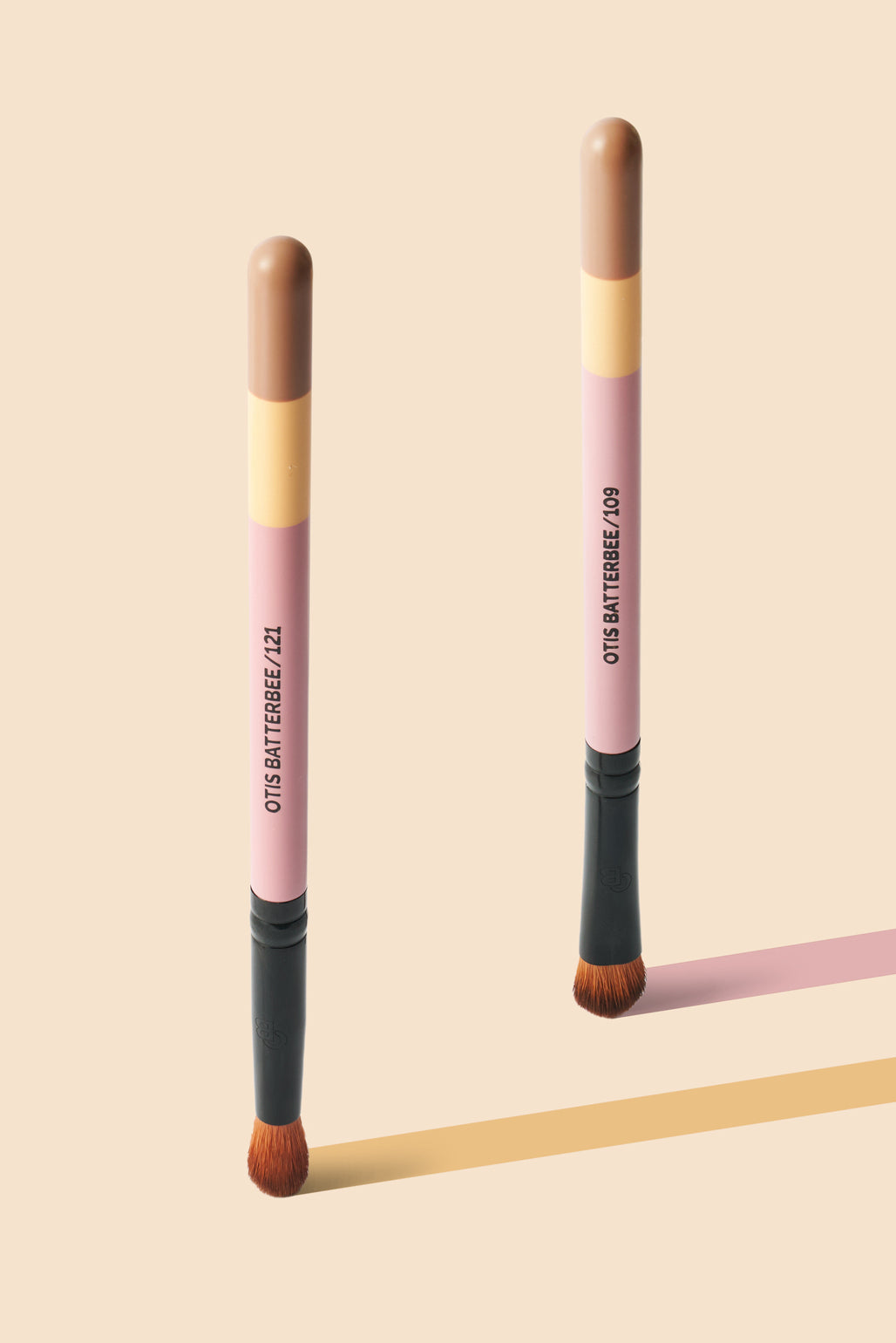 Two Otis Batterbee makeup brushes standing upright on a warm beige background, with one casting a pink shadow and the other casting a yellow shadow. The brushes have pink handles, tan and beige sections, black ferrules, and brown bristles.