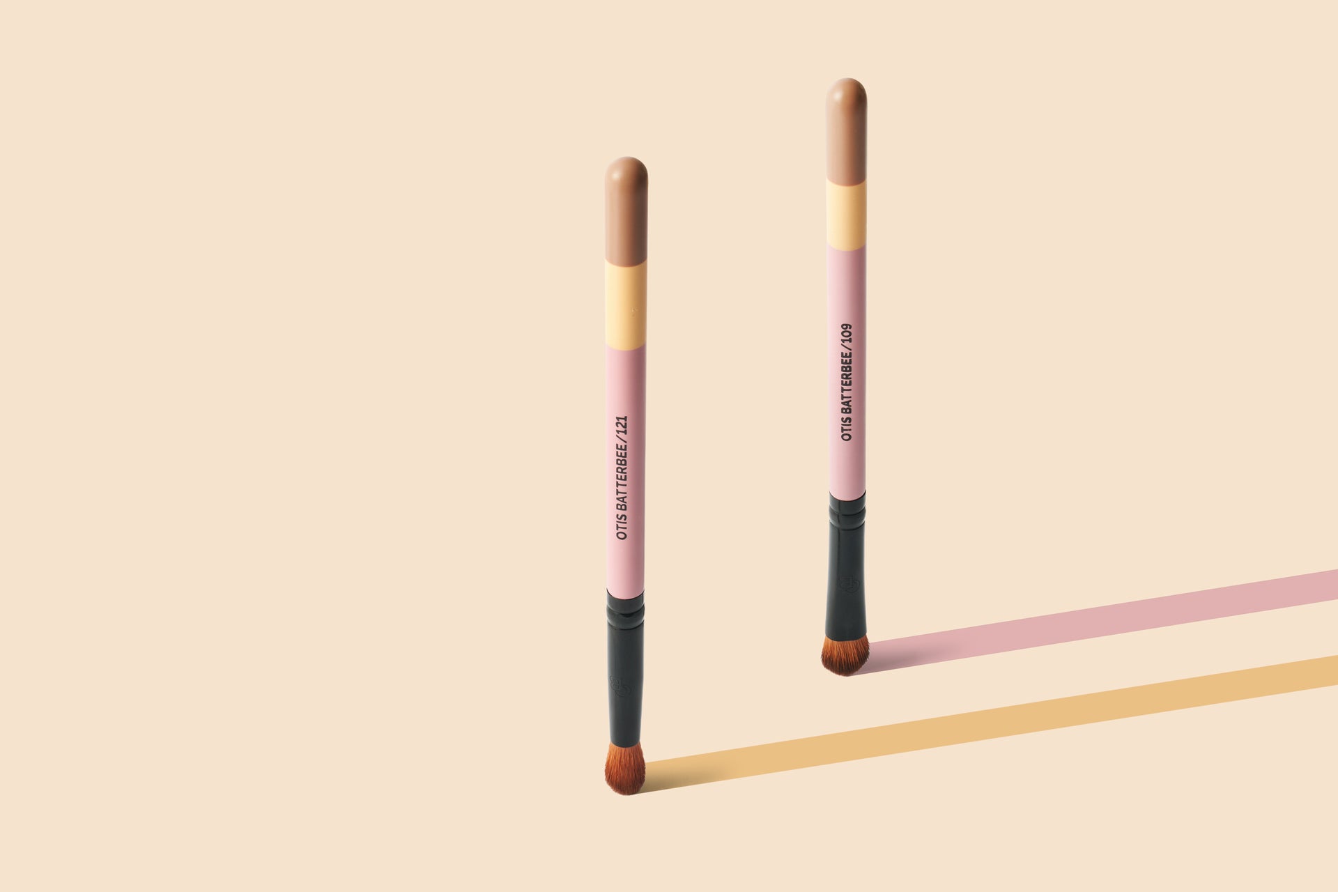 Two Otis Batterbee makeup brushes standing upright on a warm beige background, with one casting a pink shadow and the other casting a yellow shadow. The brushes have pink handles, tan and beige sections, black ferrules, and brown bristles.