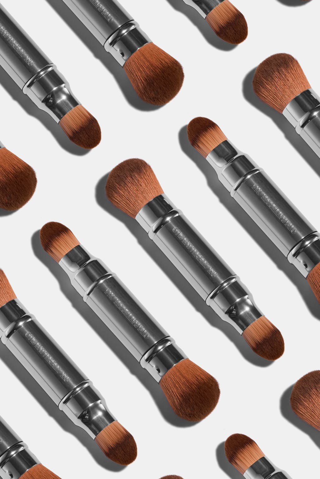 A repeating pattern of Otis Batterbee makeup brushes with silver metal handles and brown bristles, arranged diagonally on a light gray background.