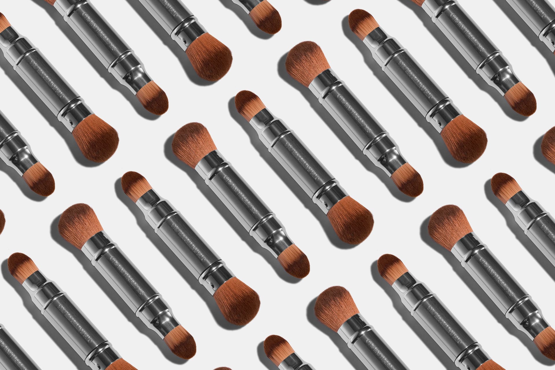 A repeating pattern of Otis Batterbee makeup brushes with silver metal handles and brown bristles, arranged diagonally on a light gray background.