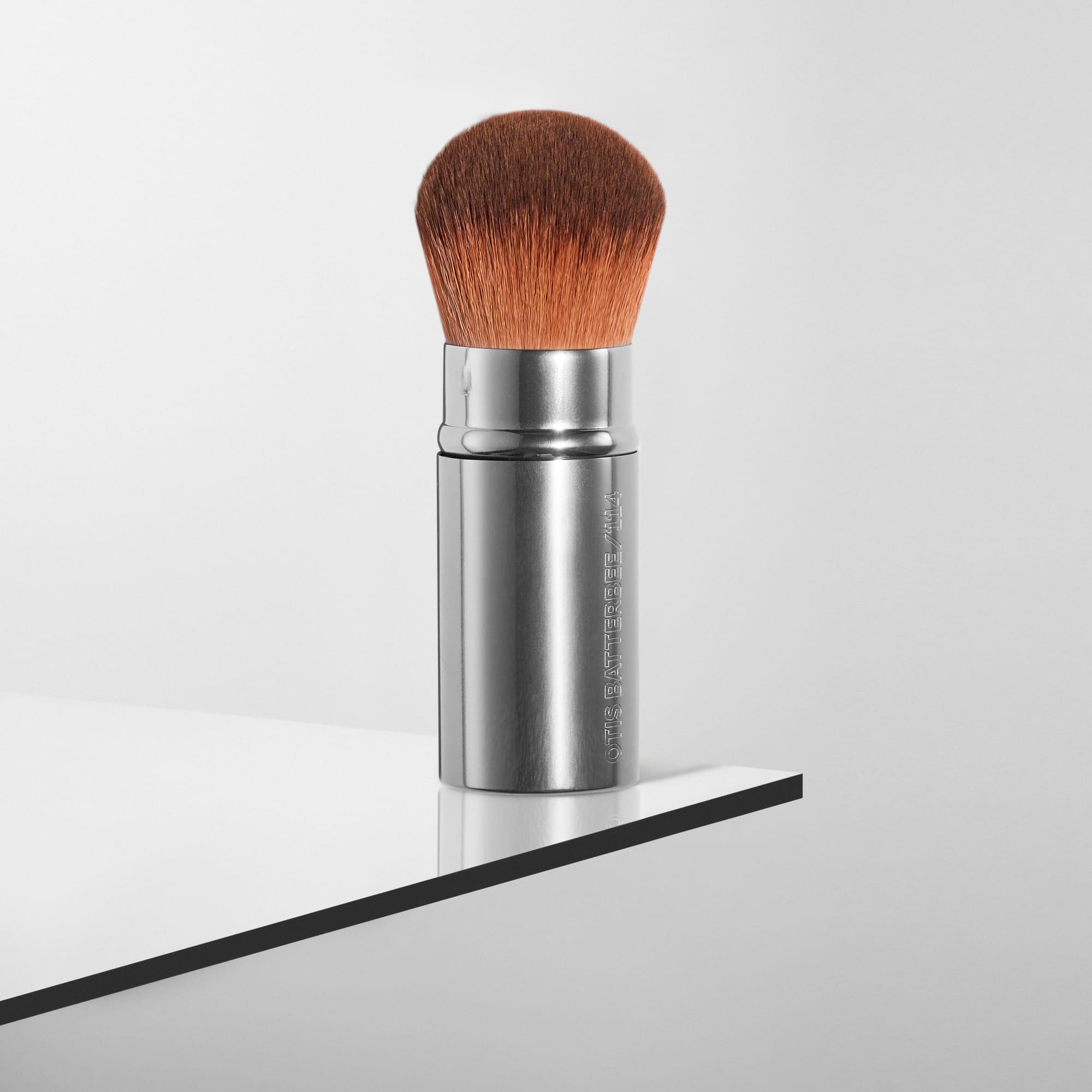 Kabuki makeup brush by Otis Batterbee with nylon bristle head and metal handle on a reflective surface